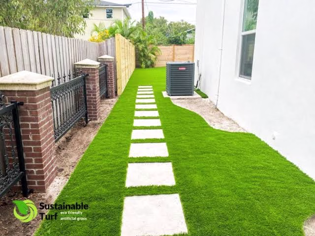 A side yard walkway with square concrete pavers set in lush synthetic grass, showcasing clean and low-maintenance Artificial Turf For Homes.