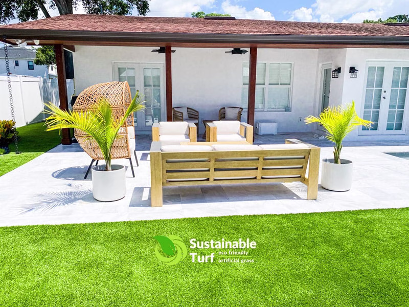 A modern covered patio with outdoor lounge furniture and potted palm trees bordering a vibrant synthetic lawn, showcasing premium Artificial Turf For Homes.